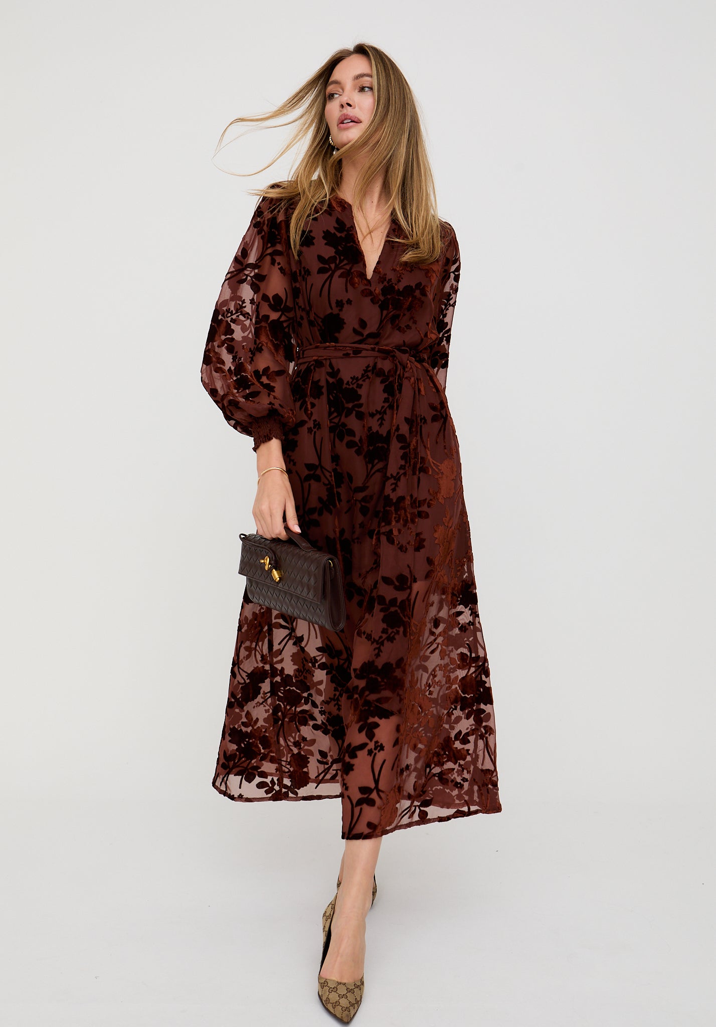 Woman wearing a brown floral dress holding a clutch against a white background