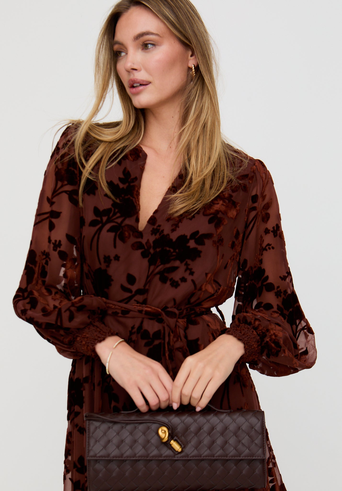 Woman wearing a brown floral dress holding a black handbag with a gold clasp on a white background