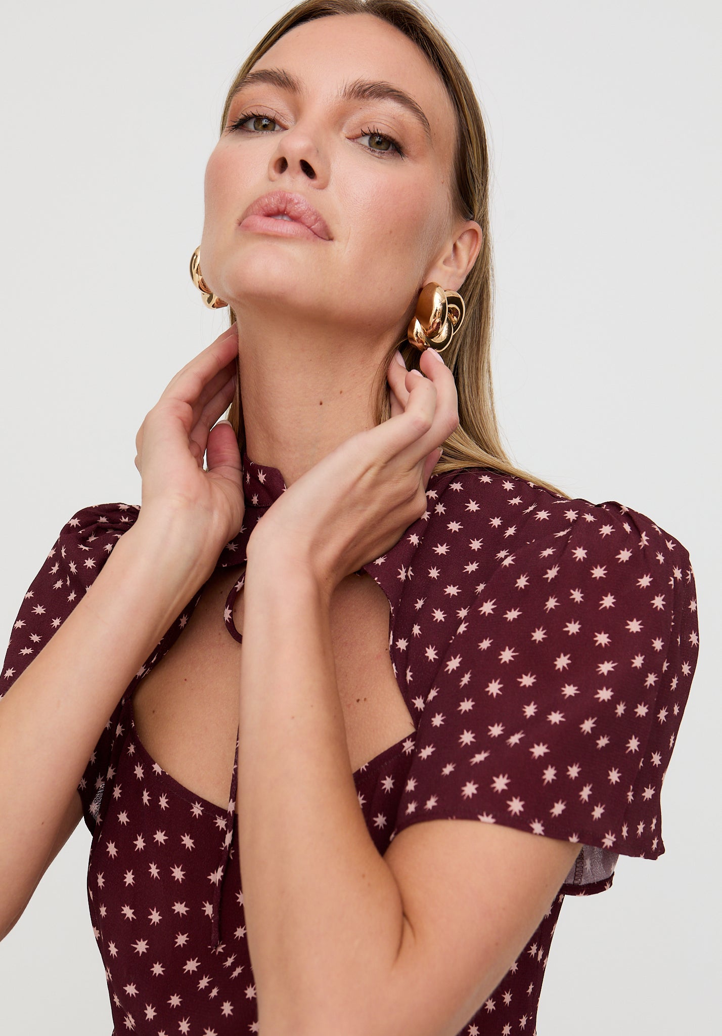 Woman wearing a burgundy top with star pattern and gold earrings on a plain background