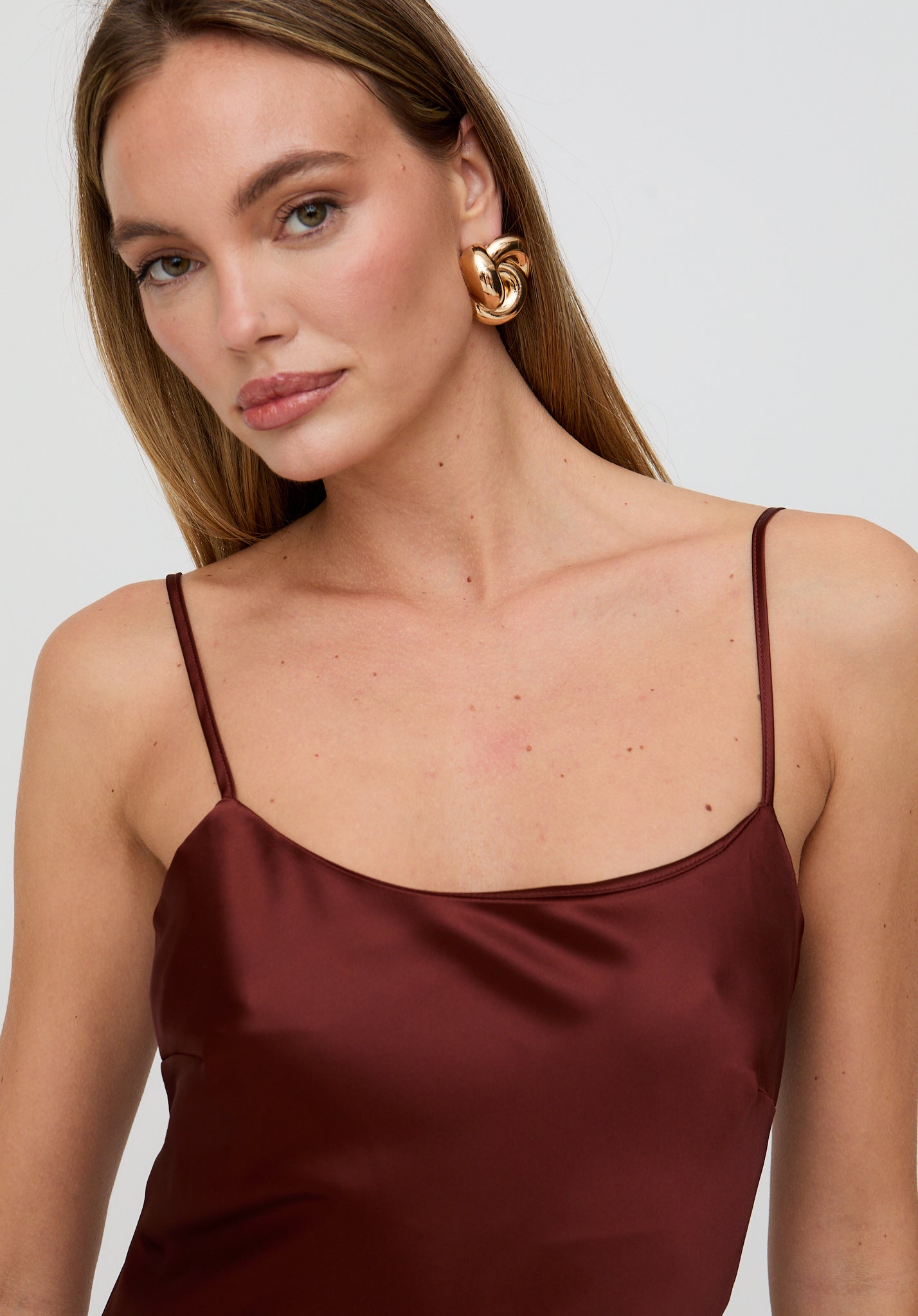 Woman wearing a burgundy dress with thin straps and gold earrings on a white background