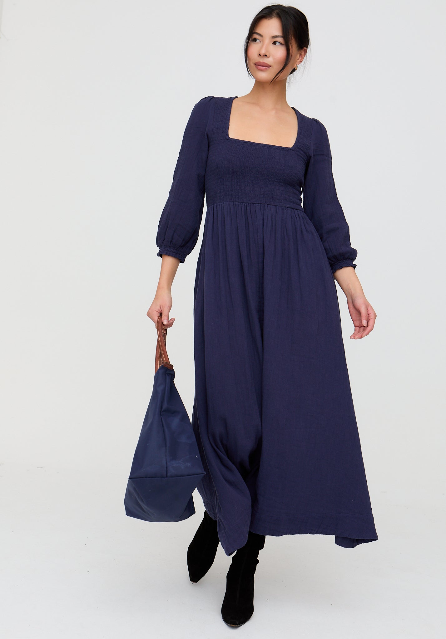 Woman wearing a long navy dress holding a matching bag against a white background