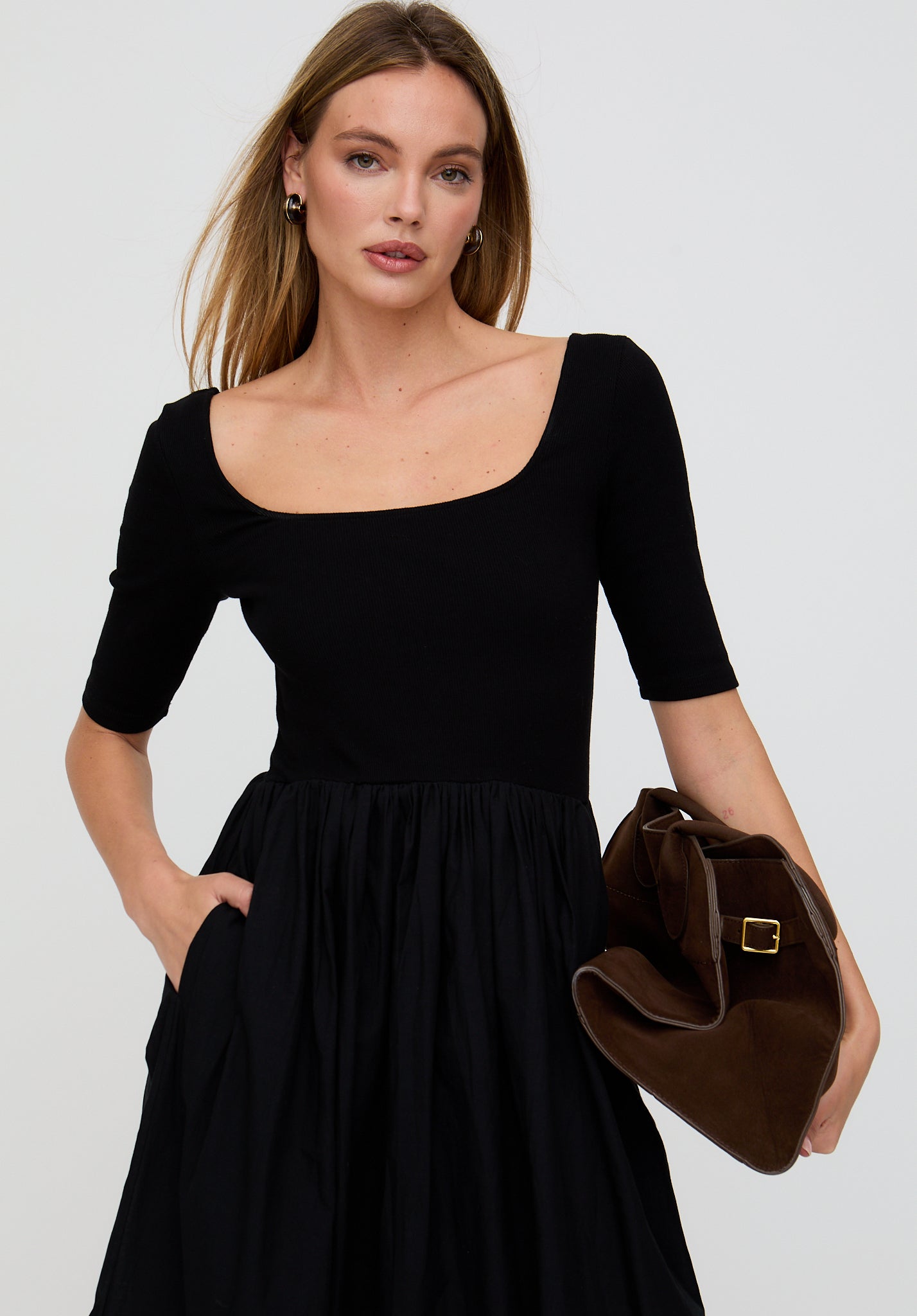Woman wearing a black dress holding a brown handbag against a plain background
