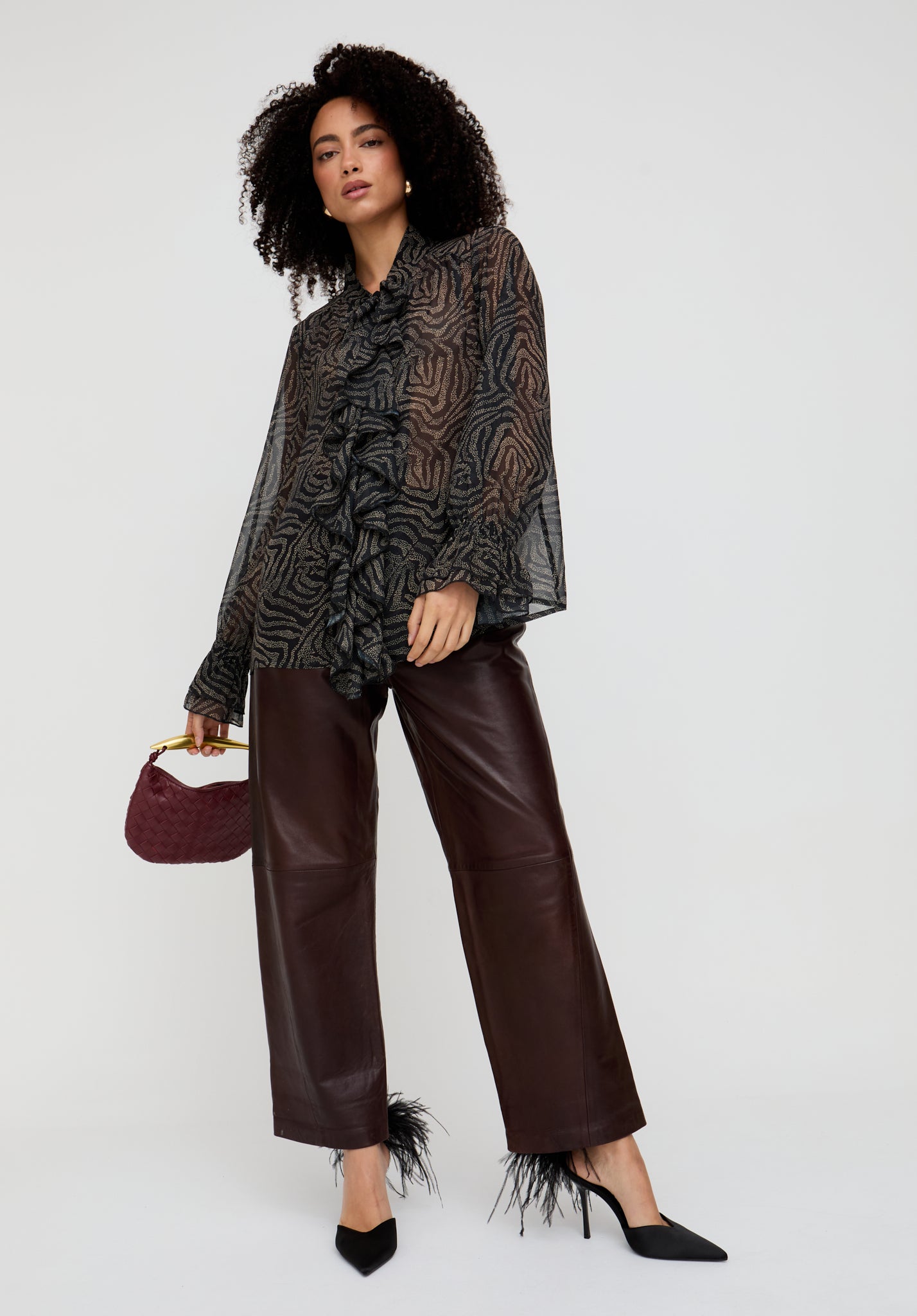 Woman wearing a patterned blouse and leather pants holding a maroon handbag on a white background