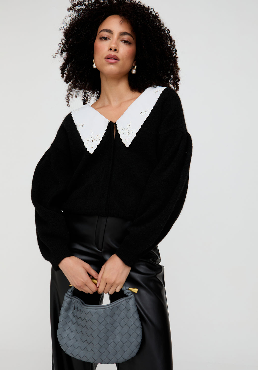 Woman wearing a black outfit with a white collar holding a gray handbag against a white background