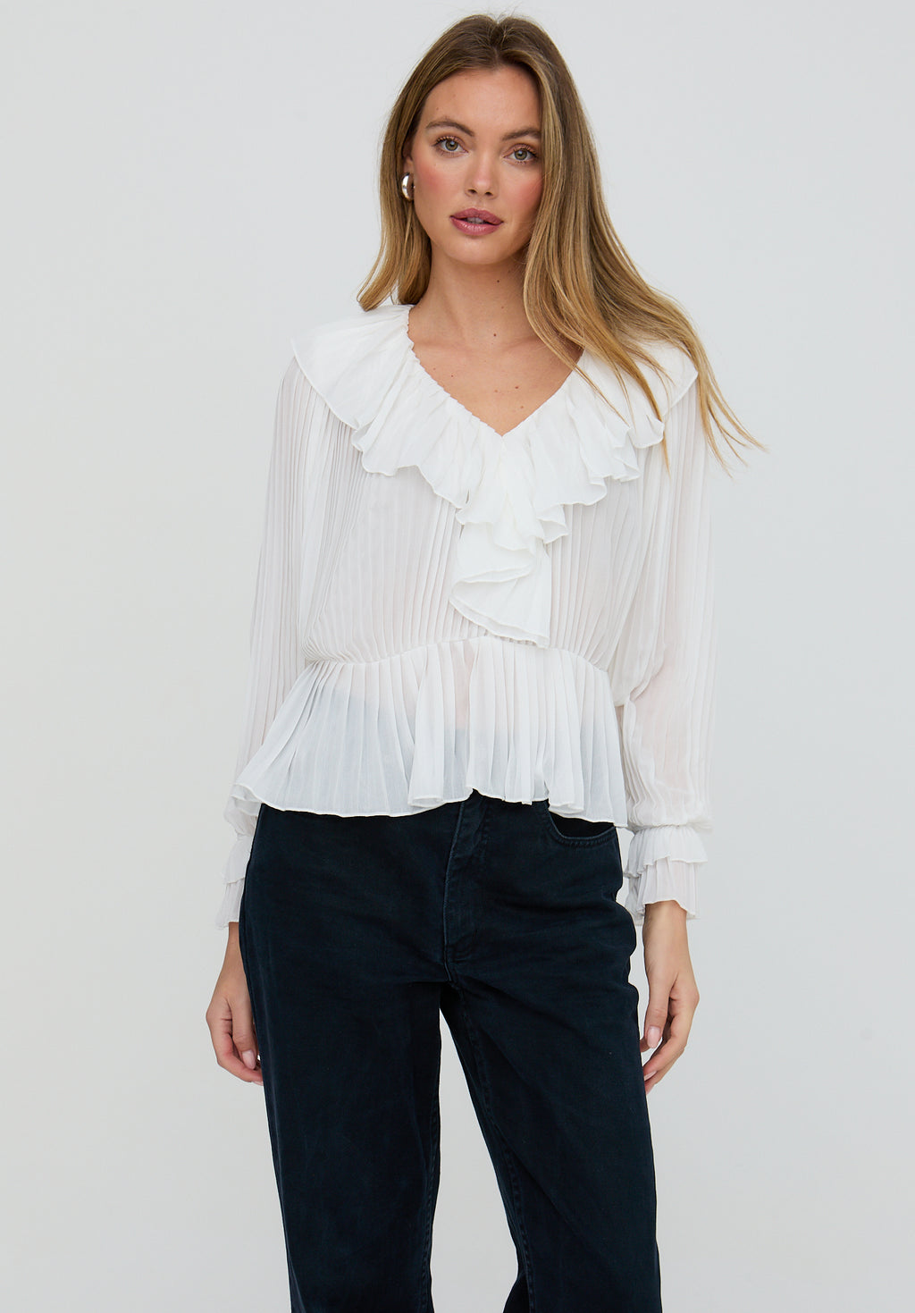 Woman wearing a white blouse with ruffles and dark pants on a plain background