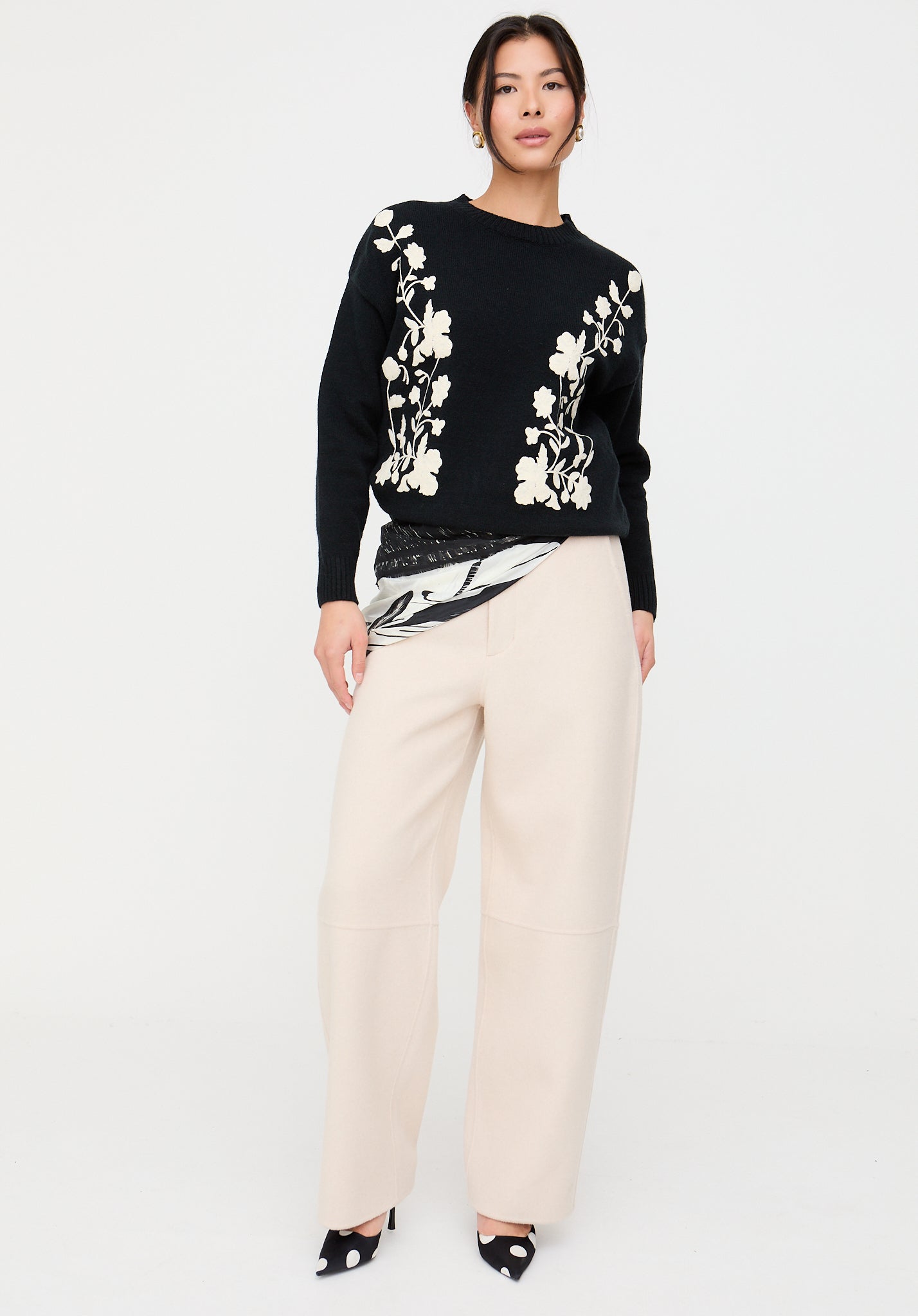 Woman wearing a black sweater with floral patterns and beige pants on a white background