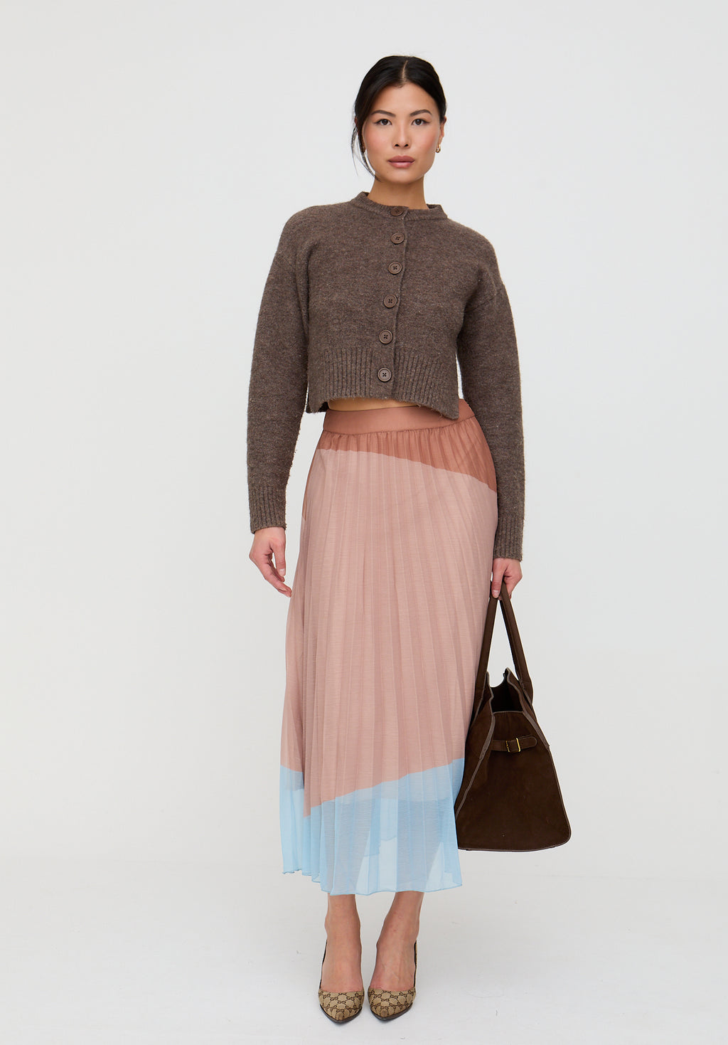 Woman wearing a brown cardigan and pleated skirt holding a brown bag on a white background